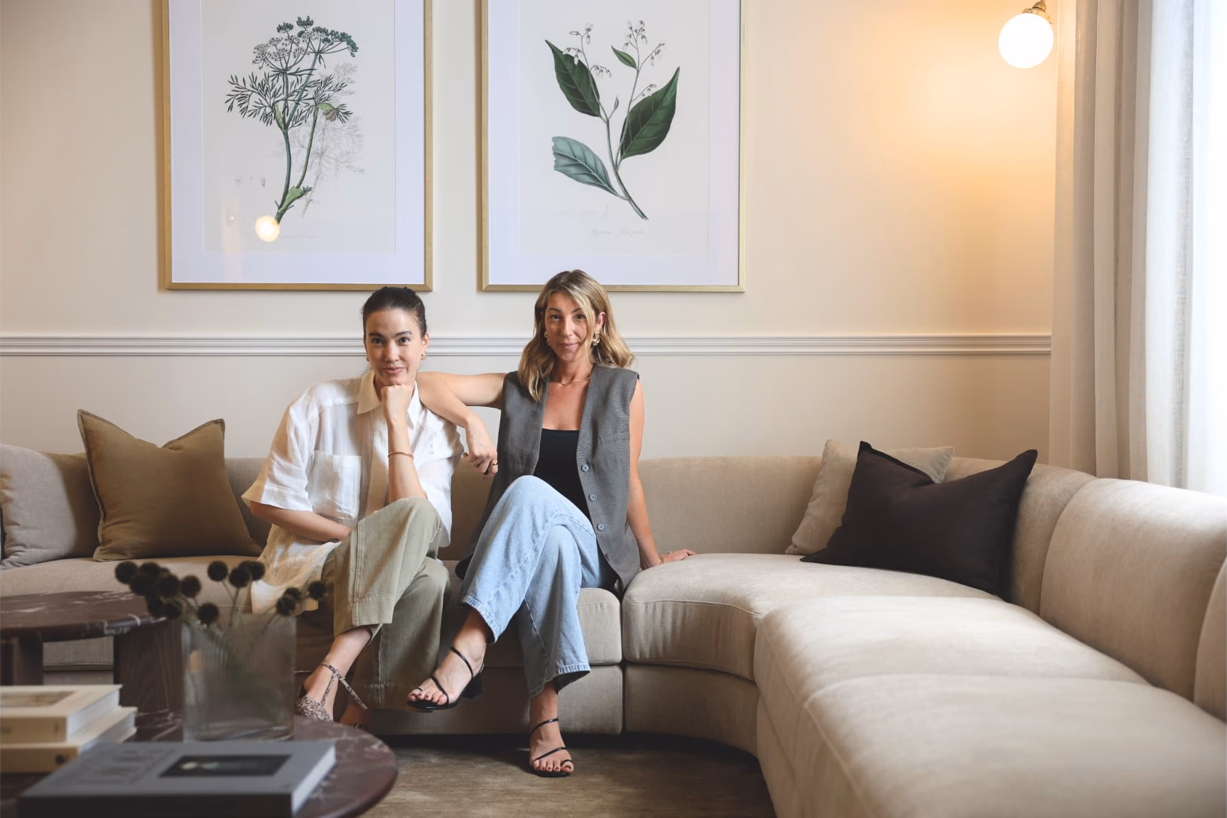 Jessica and Elisha sitting together on a curved cream sofa beneath two framed botanical prints