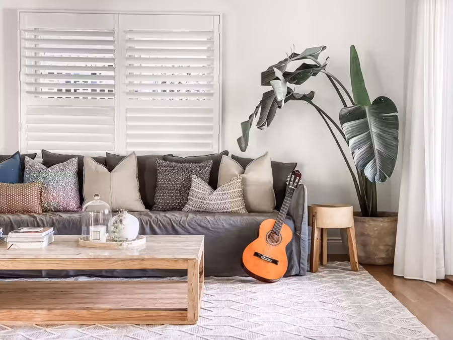 Plush grey linen slipcover sofa below plantation shutters, with a guitar and potted greenery alongside