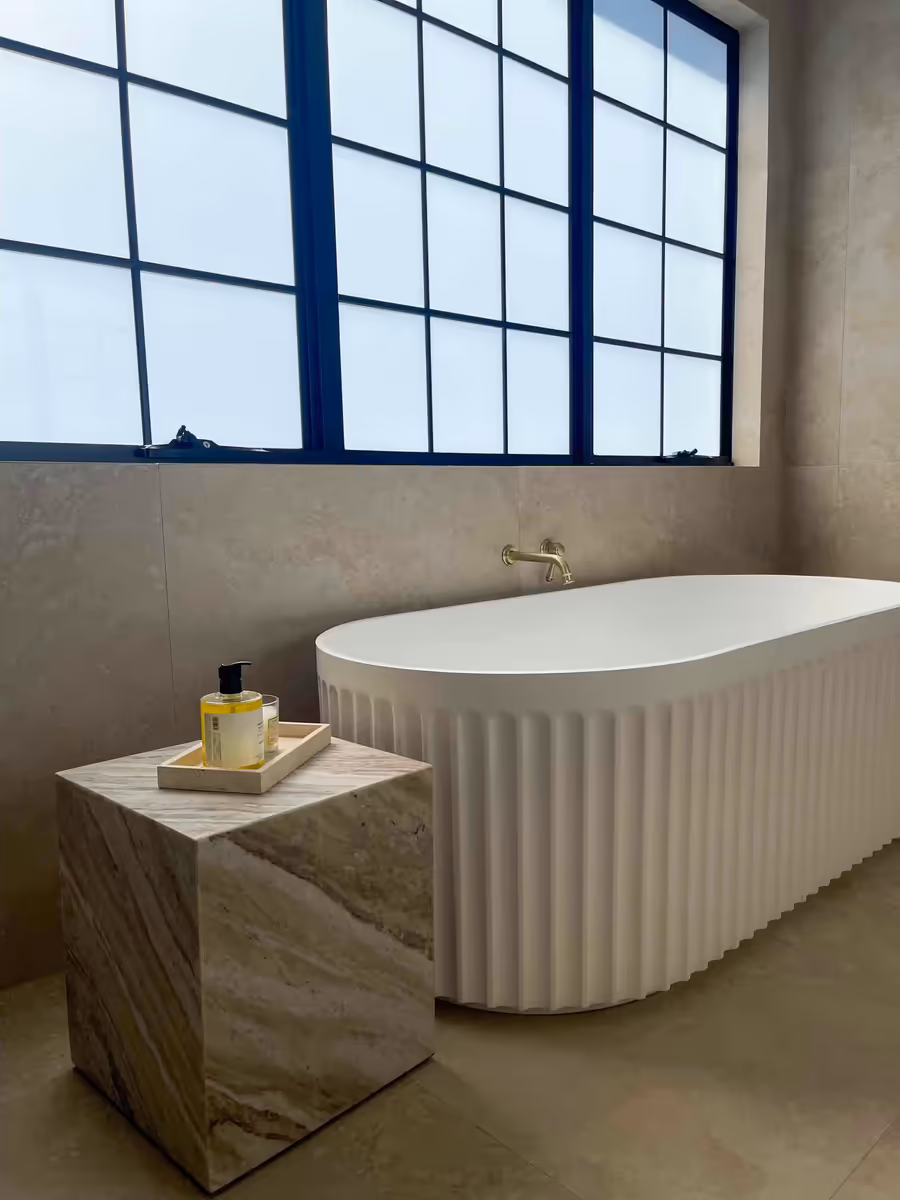 Fluted freestanding bath below a black steel window, with a marble side table