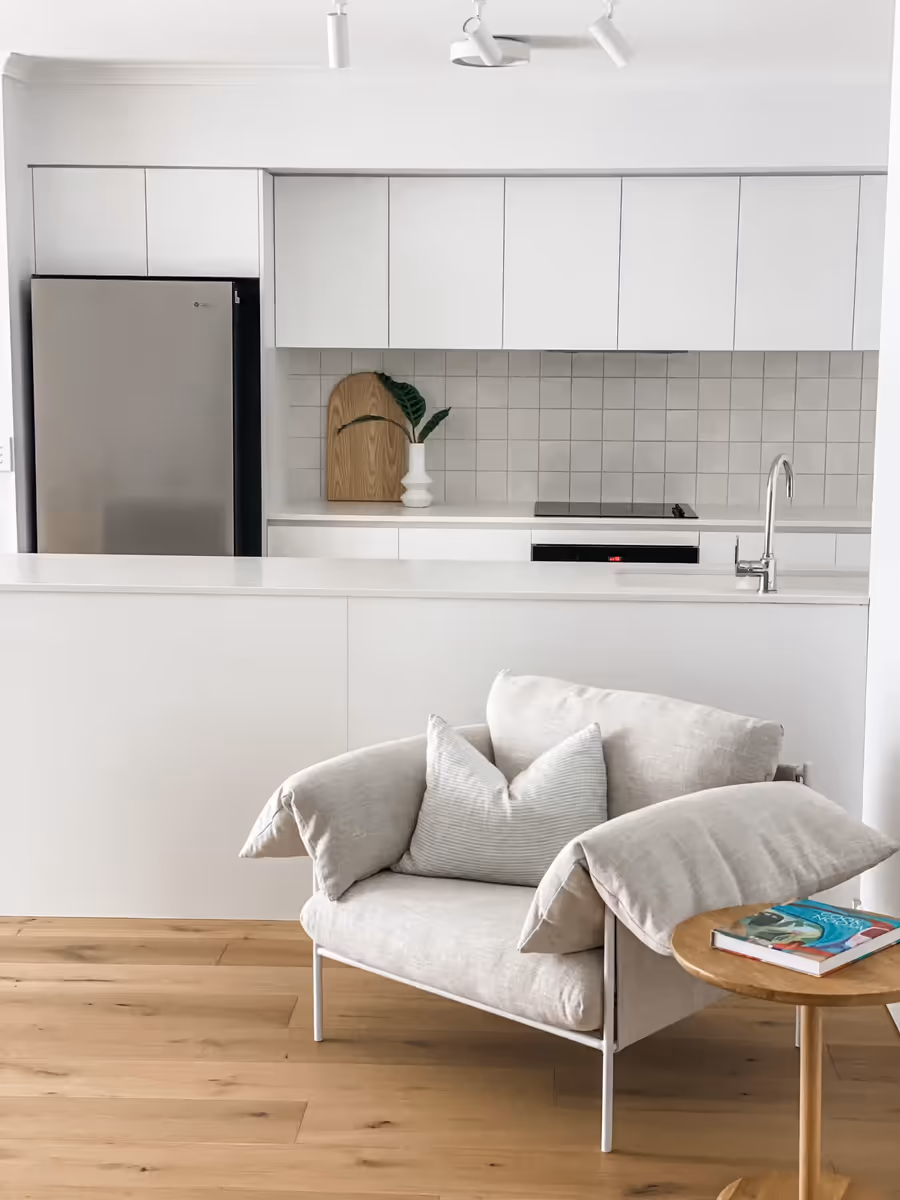 Linen armchair and a round timber side table in front of the white kitchen