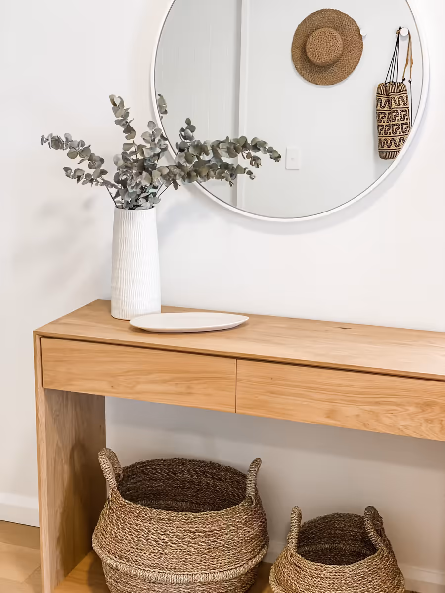 Oak console with eucalyptus and woven baskets, below a round mirror and a straw hat