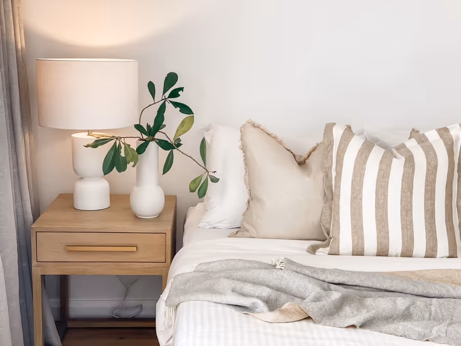 Striped and linen cushions on the bed, with a lamp and greenery on the bedside