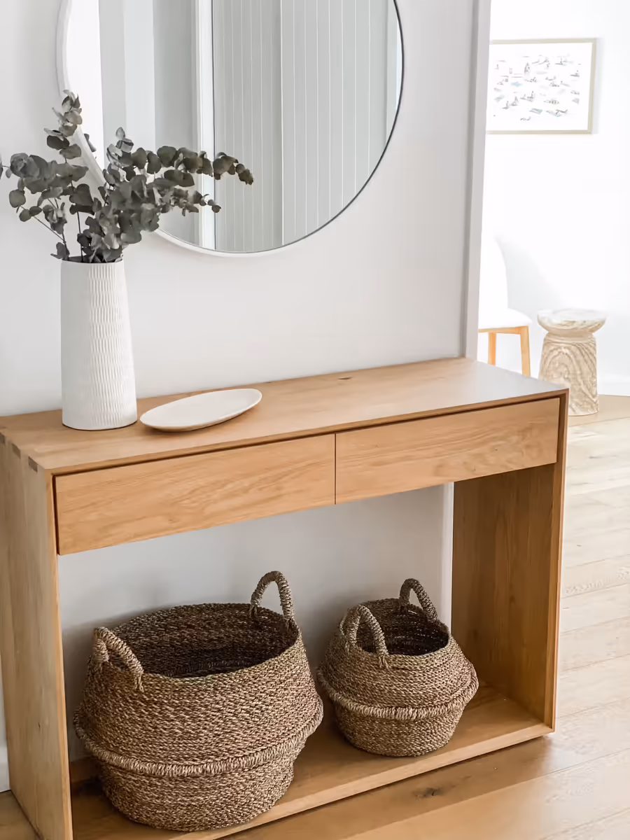 Oak console holding a vase of eucalyptus below a round mirror, baskets on the shelf