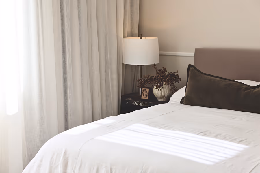 White linen bedding and an olive velvet lumbar, with a lamp on the dark bedside table